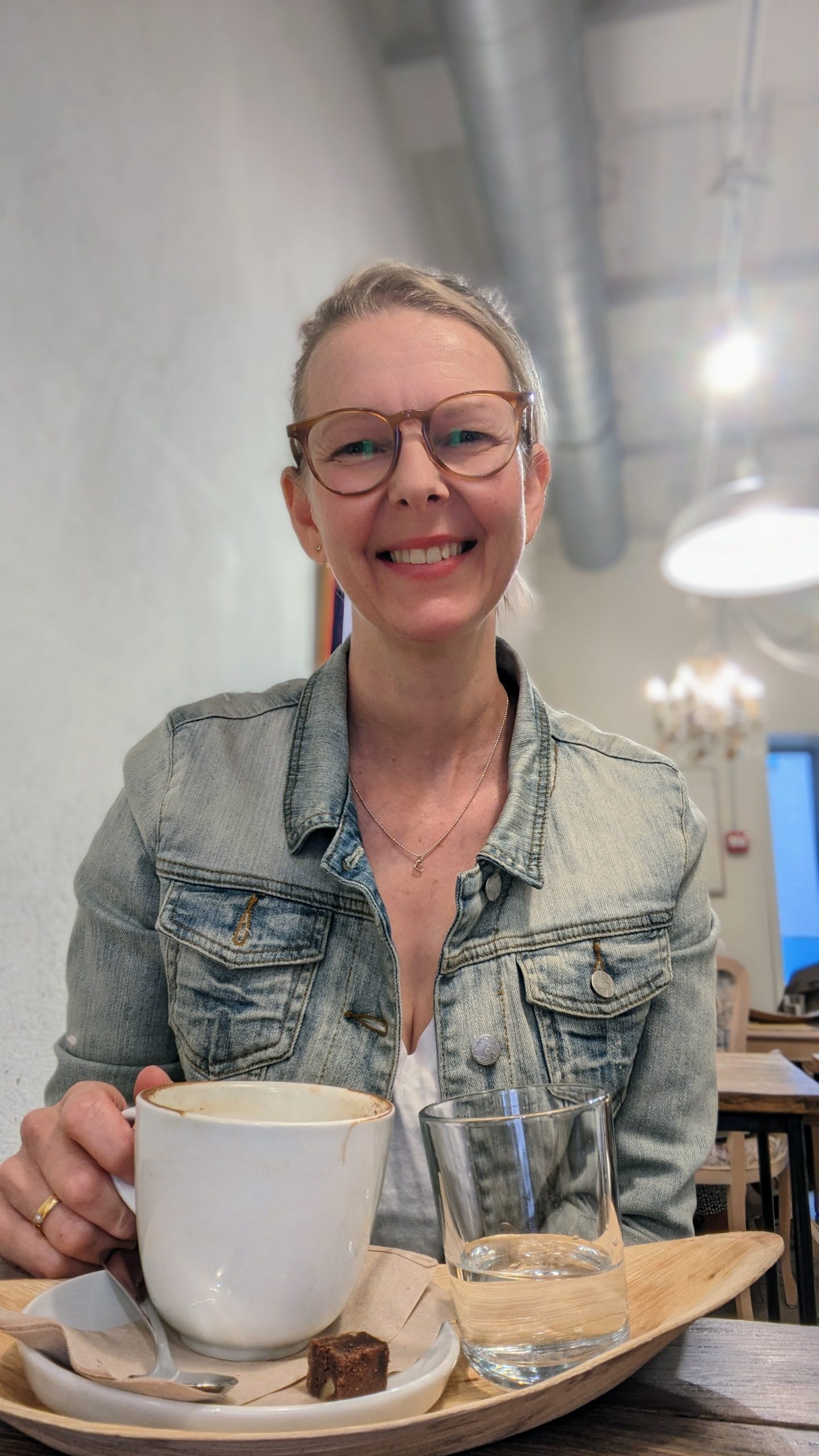 Lia Stoll, a light-skinned woman wearing seeing eye glasses and a jean jacket, holding a cup of coffee.