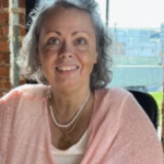 A tanned skinned woman with grey hair, wearing a pink shirt is sitting in front of a window smiling