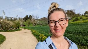 Lia Stoll, a light skinned woman with blond hair tied in a bun is wearing black seeing glasses and a blue shirt. She's standing on a field road with a green field on her right. The sky is a hazy blue with trees dotted in the background and a wooden cabin. 