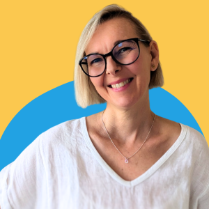Lia Stoll is standing in front of a blue and yellow wall, wearing dark-framed glasses and a white shirt is smiling at you.