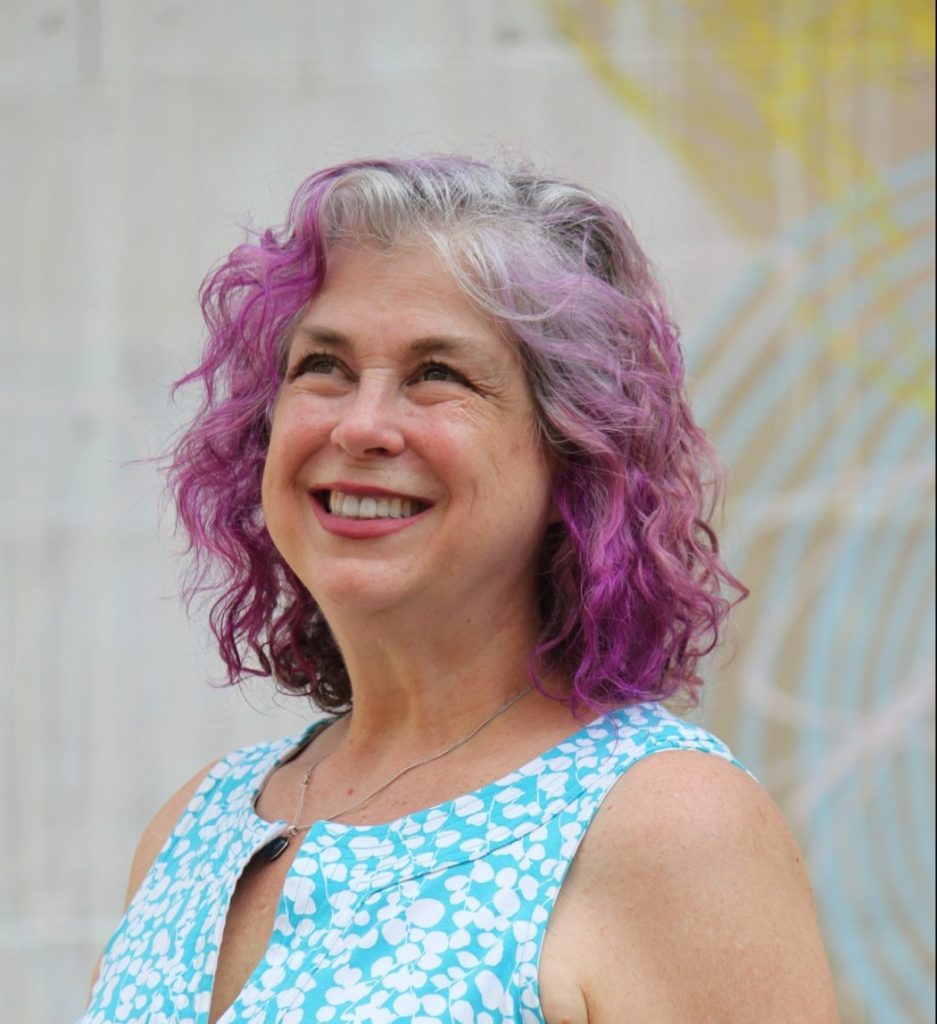 Debra Ruh, a light-skinned woman with purple hair wearing a blue and white dress is smiling