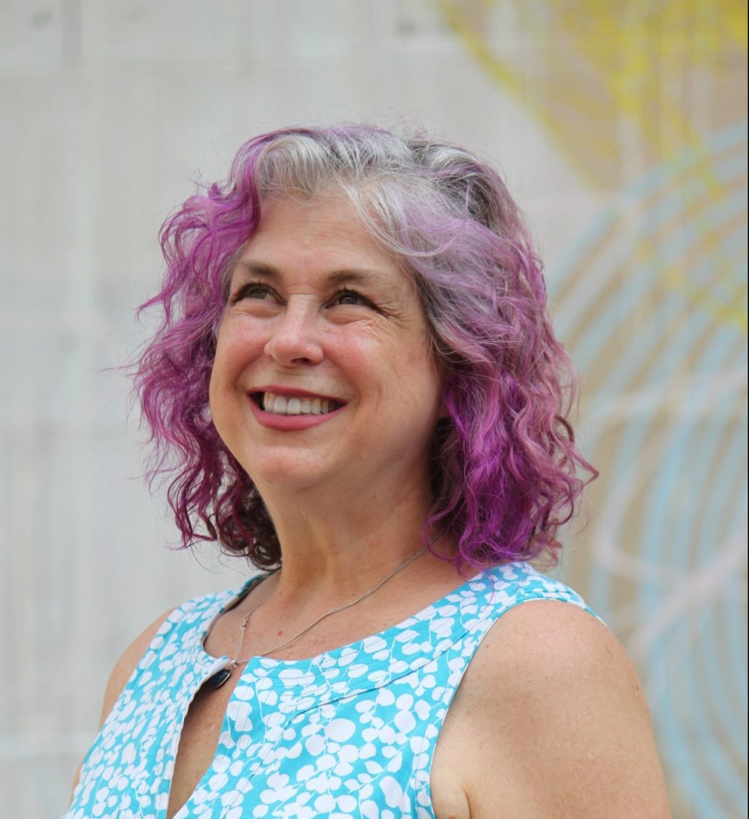 Debra Ruh, a light-skinned woman with purple hair wearing a blue and white dress is smiling
