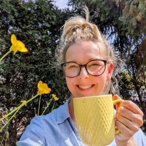 Lia Stoll in her garden, wearing dark-framed glasses and a blue shirt is holding a yellow coffee cup smiling 