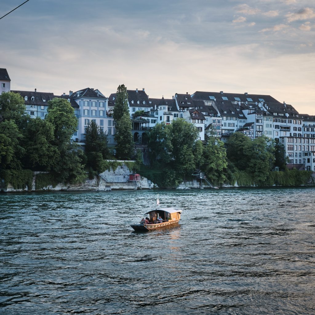 Basel city on the banks of the river Rhein during dusk. There's a small ferry boat crossing. 