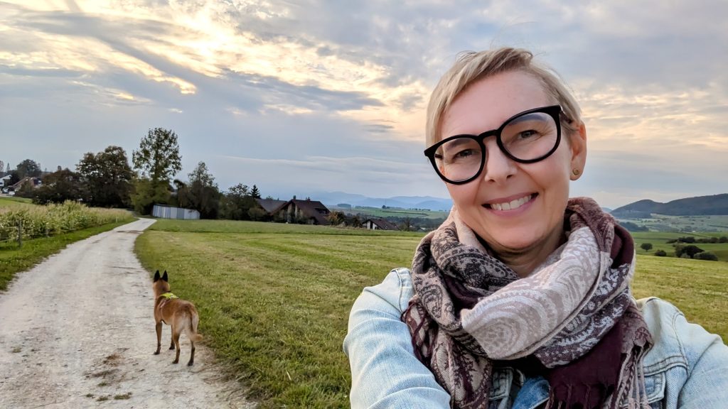 Disability Writer, Lia Stoll, a light-skinned woman 
with short blond hair is standing on a field road wearing dark-framed glasses, a jean jacket and a scarf. 